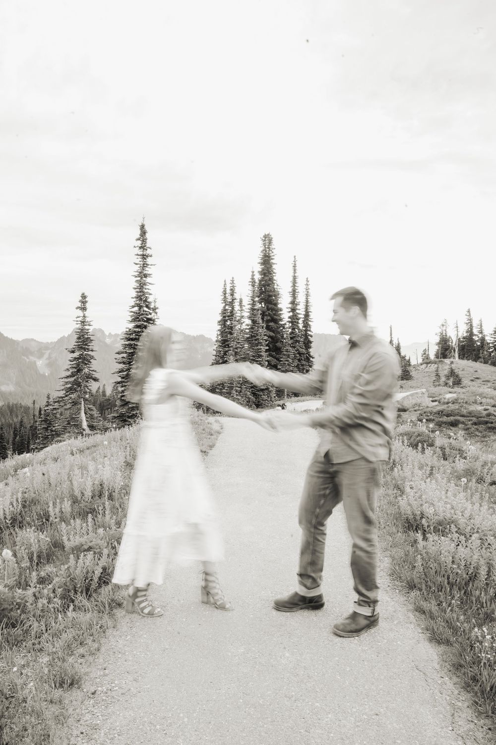 A Engaged couple dances together on a mountain trail surrounded by evergreen trees and scenic mountain views in black and white.