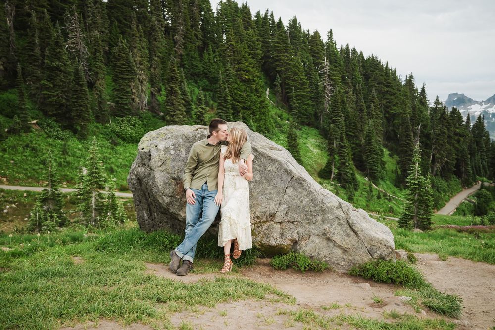An Engaged couple on a scenic hiking trail winds through a dense evergreen forest with large boulders scattered along the mountainside path.