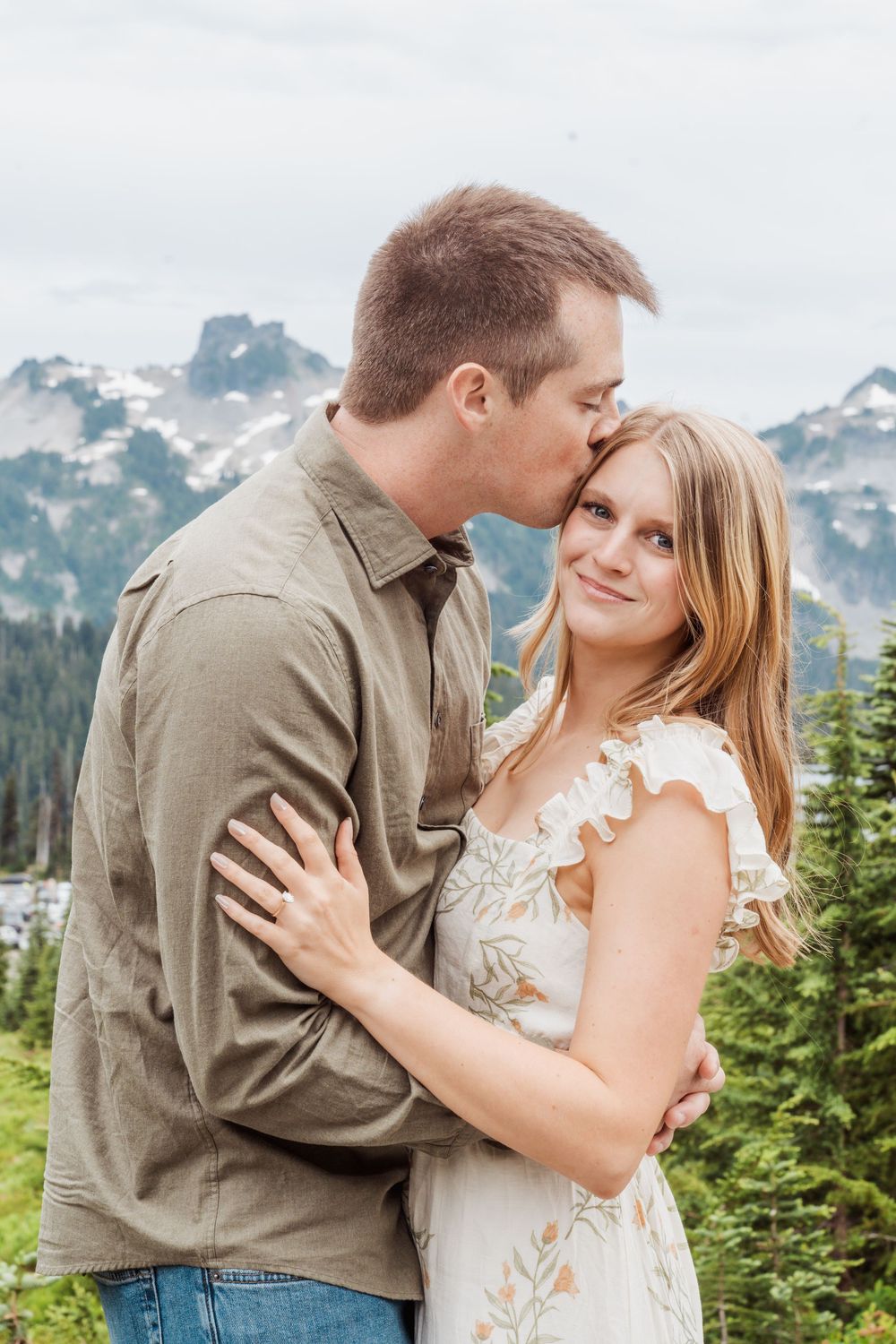 An Engaged couple embraces against a scenic mountain backdrop with snow-capped peaks and evergreen forest.