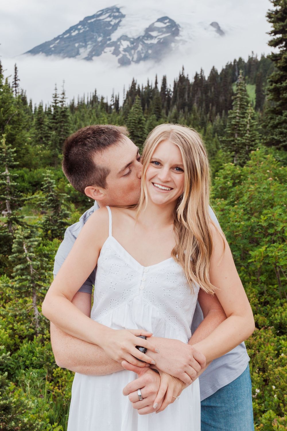 A Engaged couple embraces in front of a misty mountain peak with evergreen forest in Mount Rainier National Park.