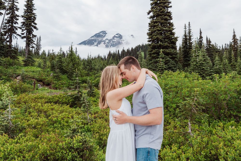 An engaged couple embraces in a meadow with Mount Rainier rising majestically in the background through evergreen trees.
