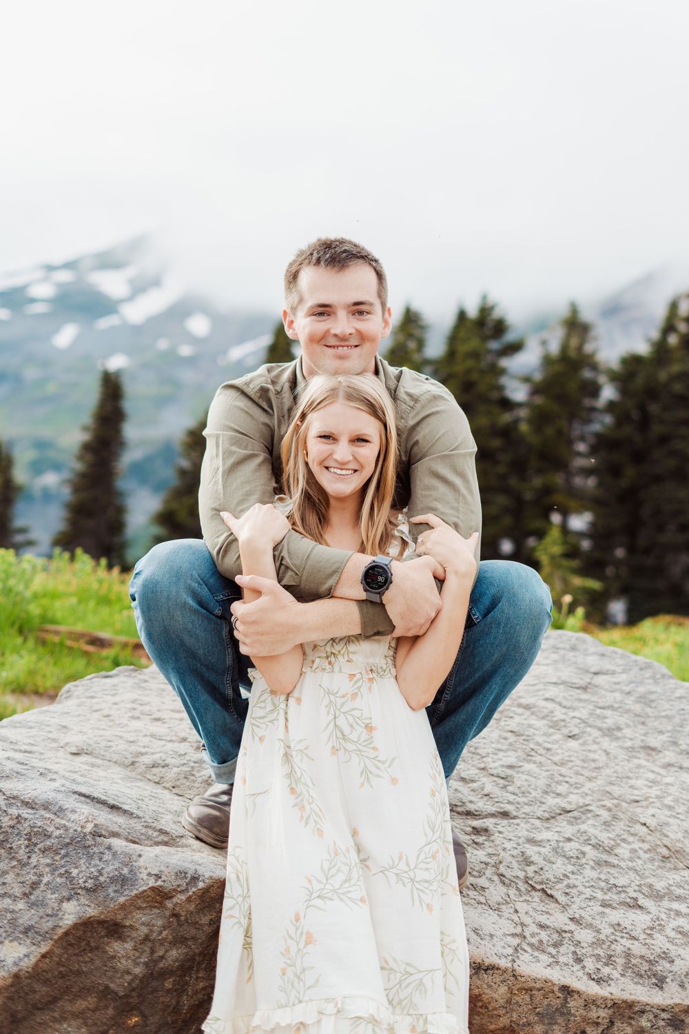 A couple shares playful moments on a rocky mountain overlook with evergreen trees and snow-capped peaks in the distance.