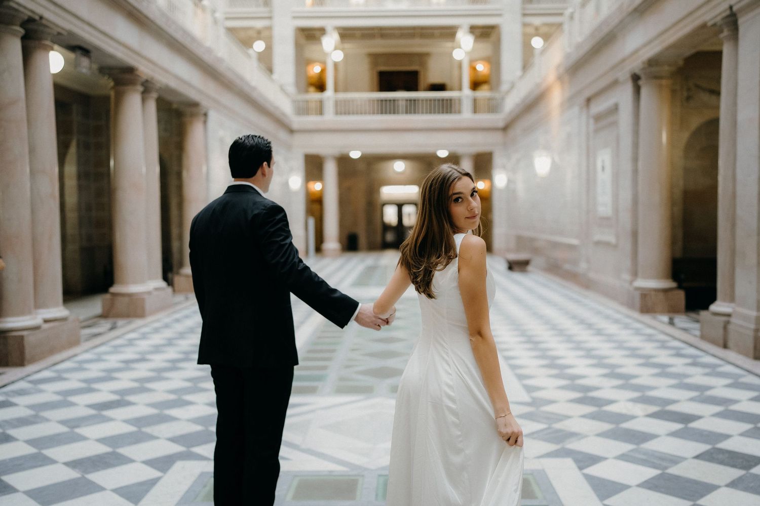 Elegant couple in formal wedding attire poses in a grand marble hallway with checkered floors and ornate columns.