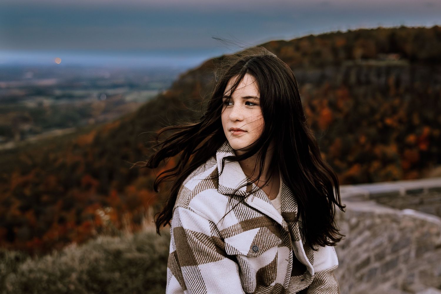 Woman in white coat stands against autumn mountain landscape with dramatic cloudy sky in background.