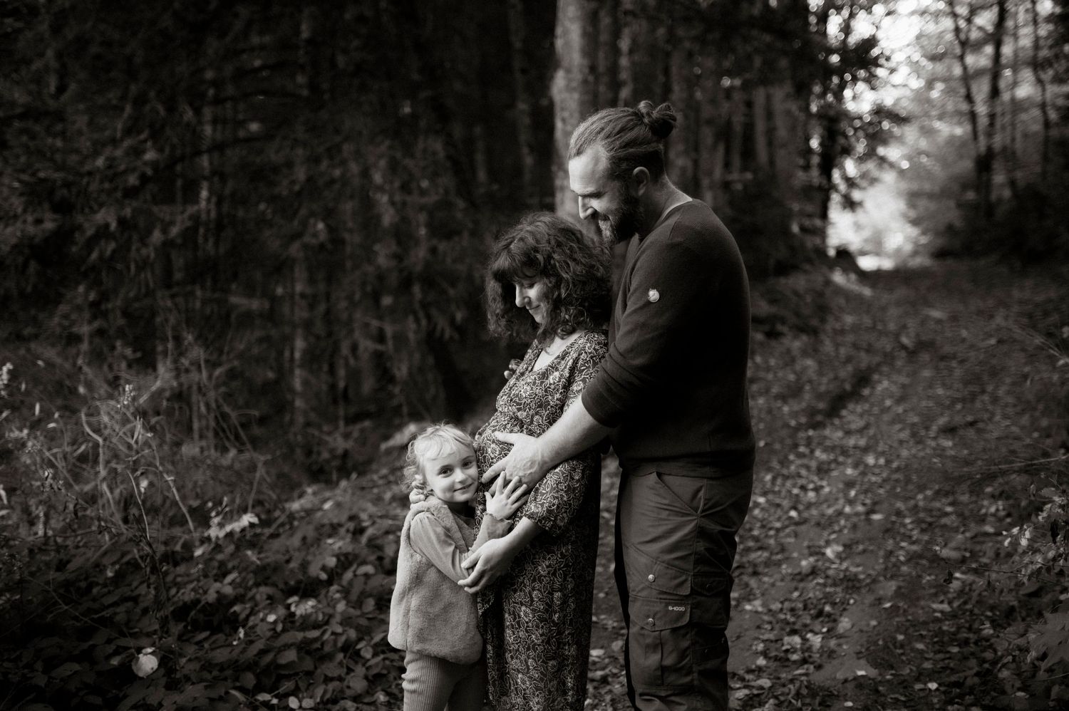 A black and white photograph shows a tender moment shared between people embracing on a wooded forest path.