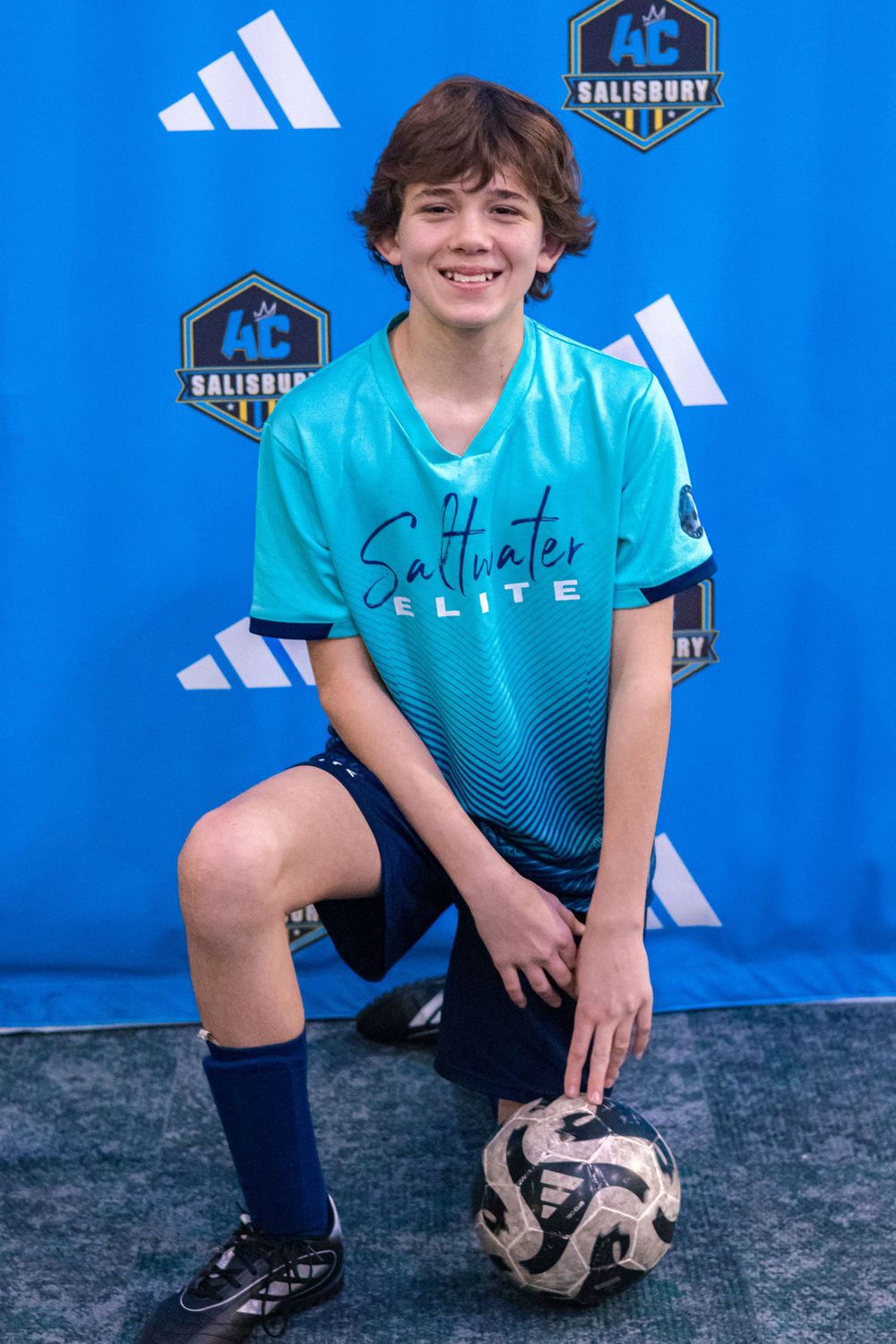 Young soccer player in turquoise jersey poses with ball against blue backdrop with Adidas and Salisbury logos.