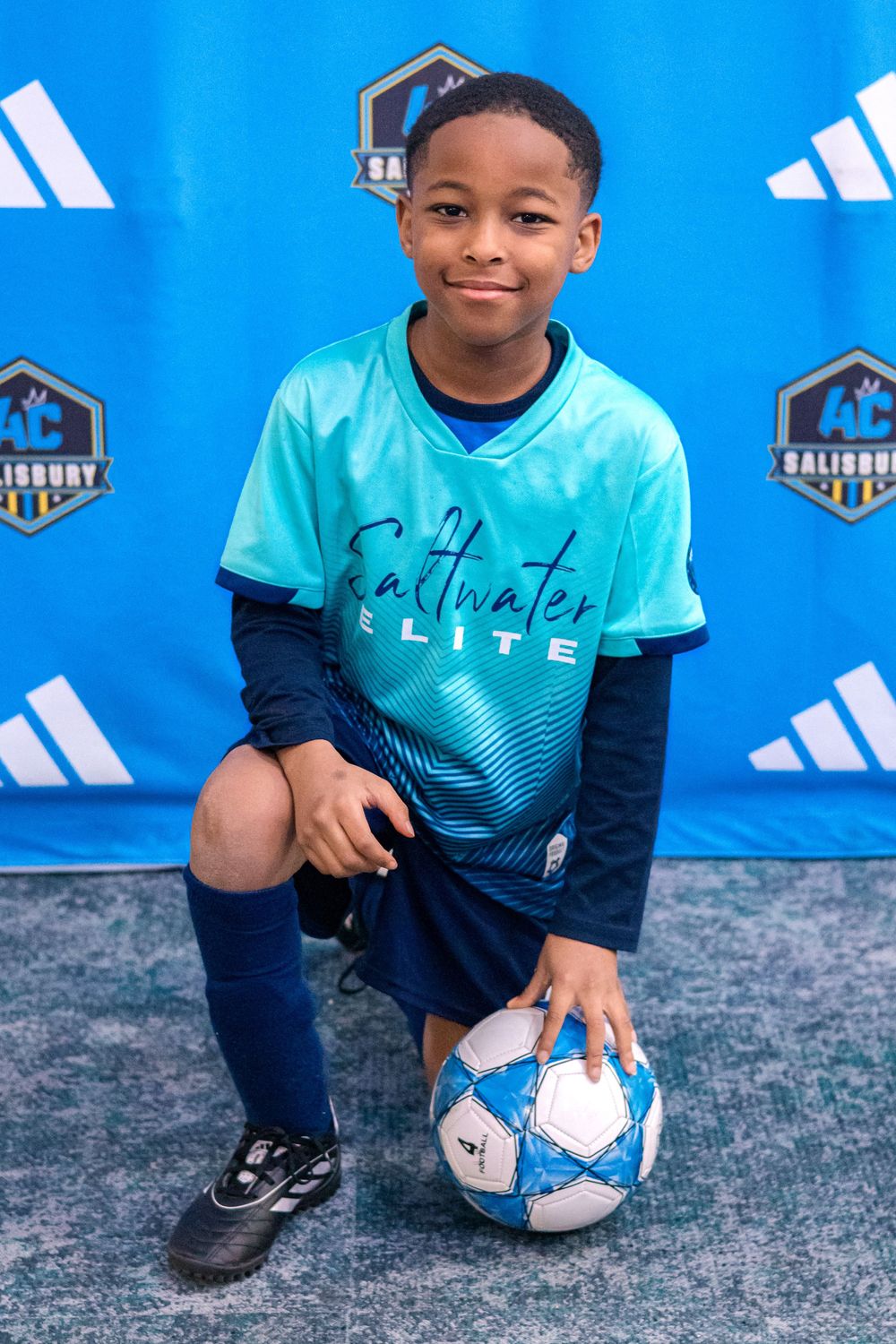 Soccer player in turquoise and navy uniform poses with white soccer ball against blue backdrop.