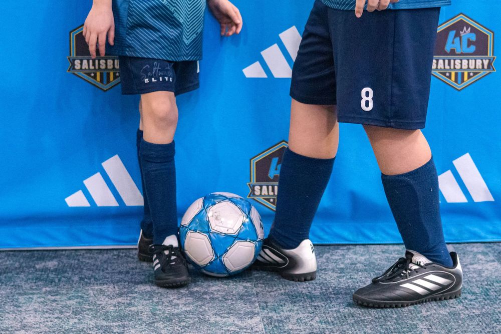 Soccer player in turquoise uniform kneels with ball on gray carpet against blue background.