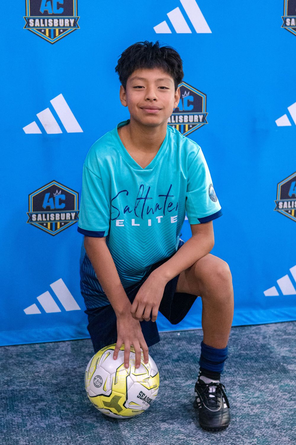 Youth athlete in turquoise Elite jersey kneels with soccer ball against blue background.