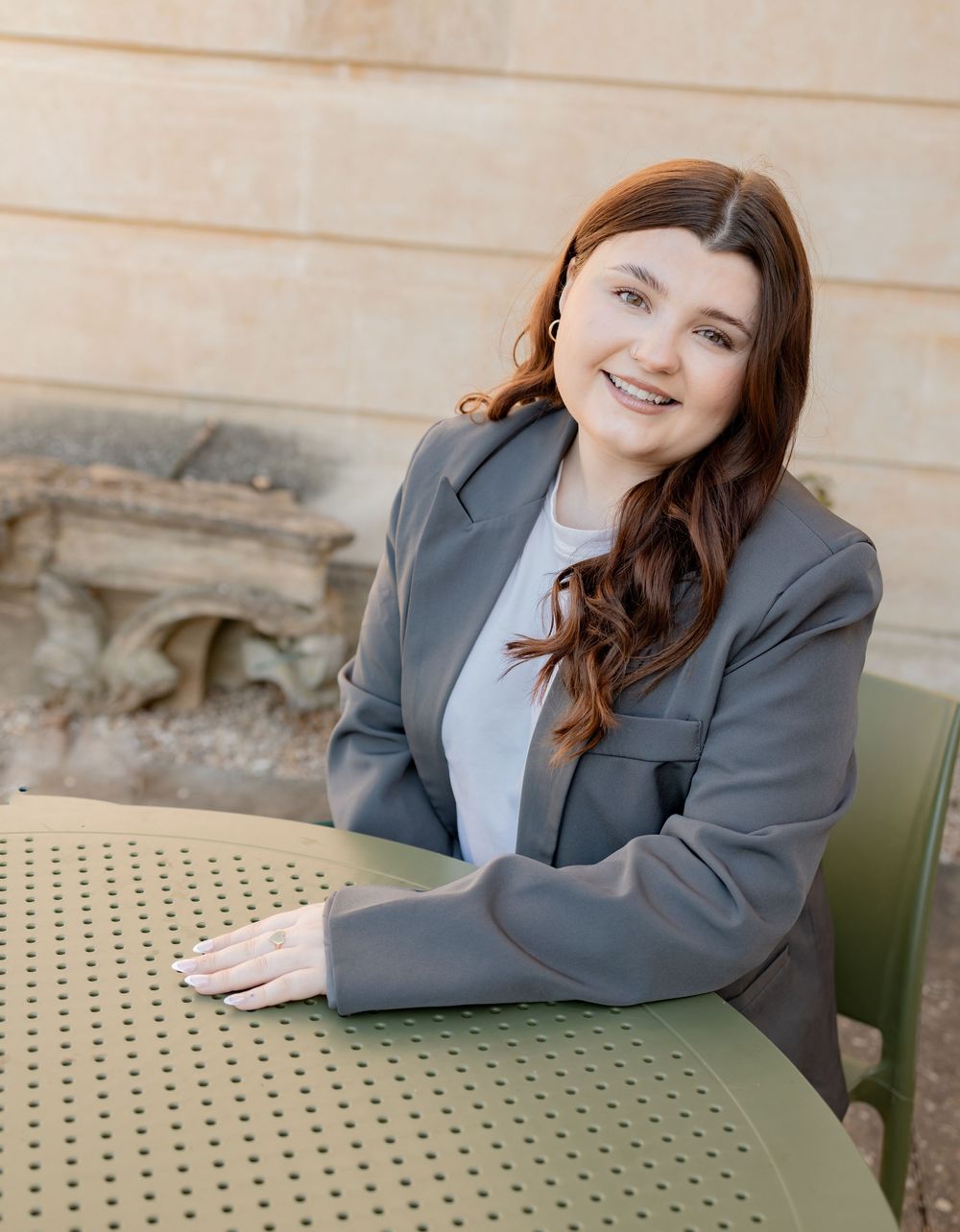 Young woman in gray blazer smiling at outdoor table with stone architecture background. Photography in Buckinghamshire.