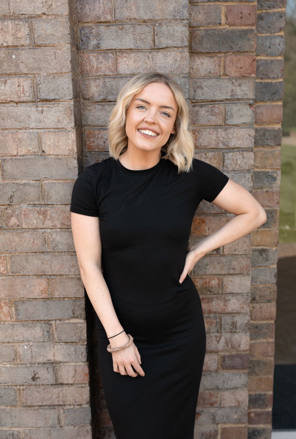 Smiling blonde woman in black fitted dress posing against rustic brick wall outdoors. Photography in Buckinghamshire.