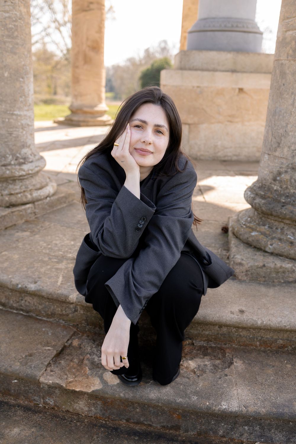 Young woman in black blazer crouching gracefully among ancient stone columns outdoors. Photography in Stowe, Buckinghamshire.