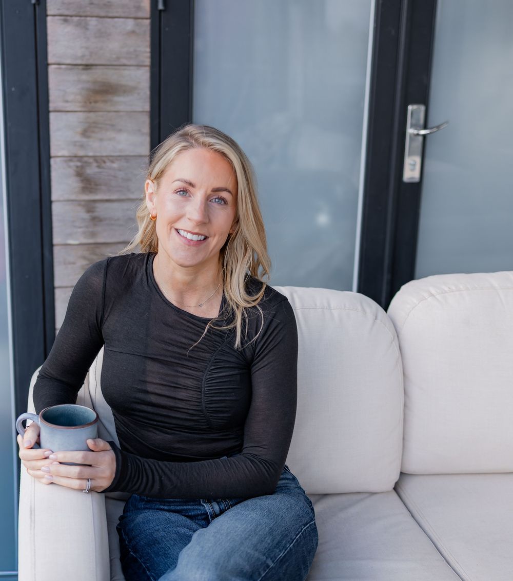 Smiling blonde woman relaxing on white outdoor sofa holding gray mug, wearing black long-sleeve top and jeans.
