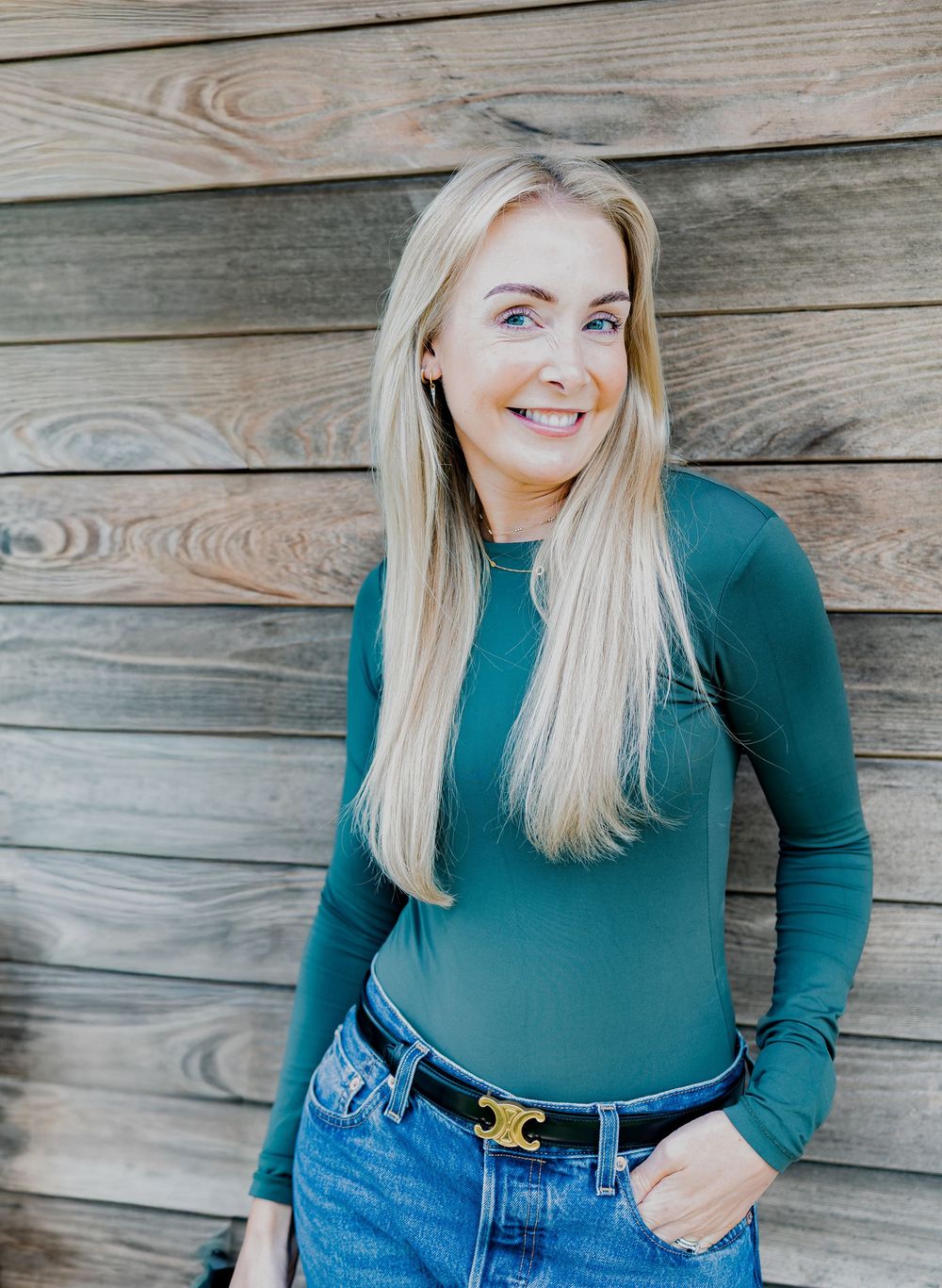 Smiling blonde woman in teal long-sleeve top and jeans poses against rustic wooden wall background.