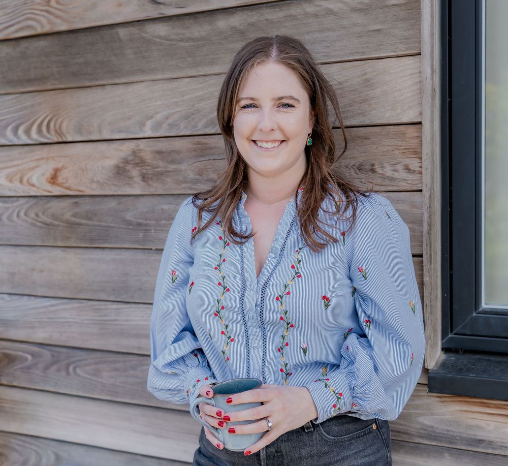 Smiling woman with brown hair wearing a floral embroidered blue striped blouse, standing against a wooden wall.
