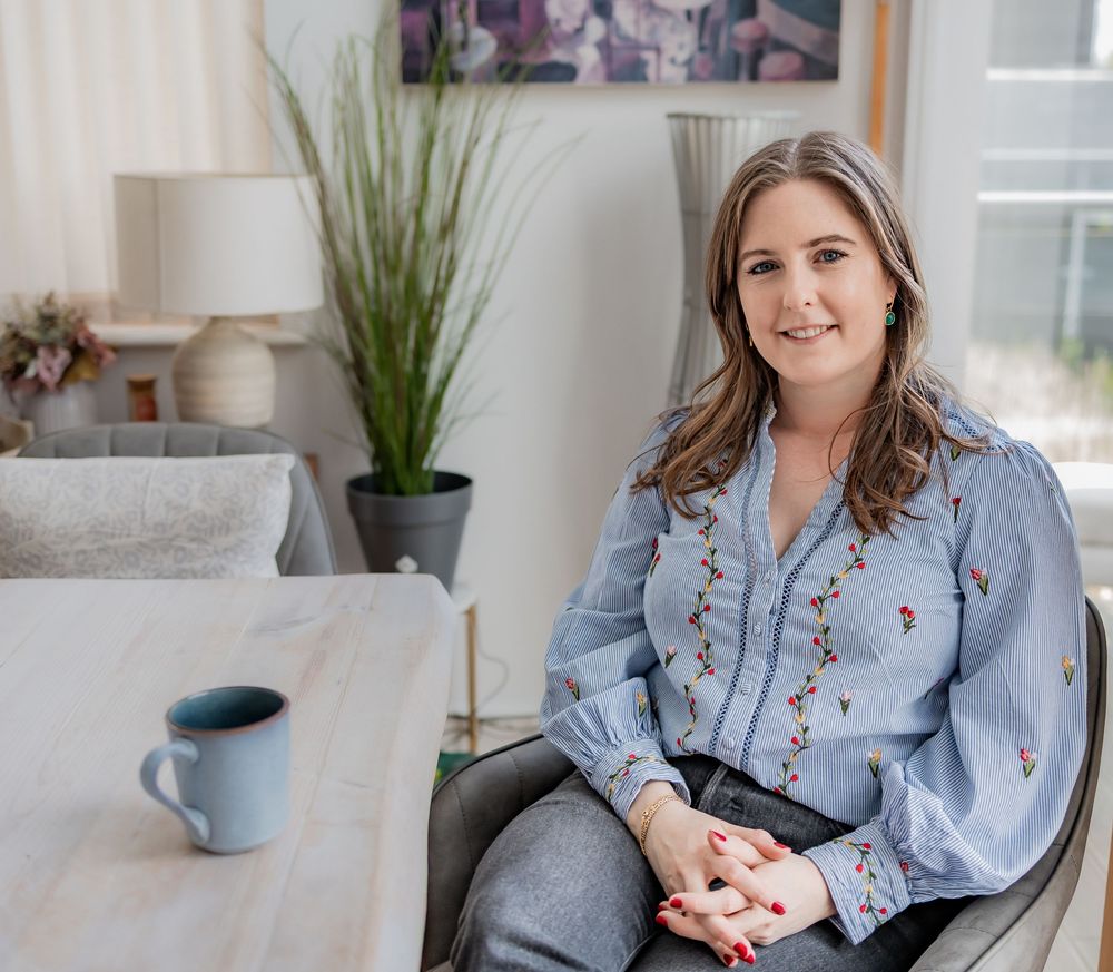 Woman in blue floral blouse sitting at a table with a coffee mug in a bright modern living room.