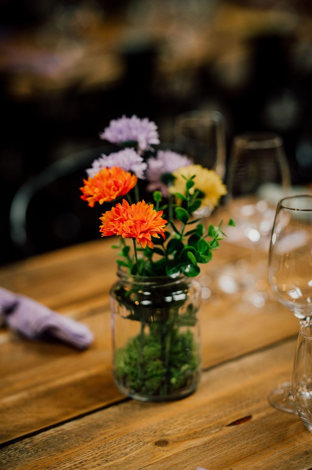 Small orange chrysanthemum flowers in glass jar vases arranged in a row on rustic wooden table surface.