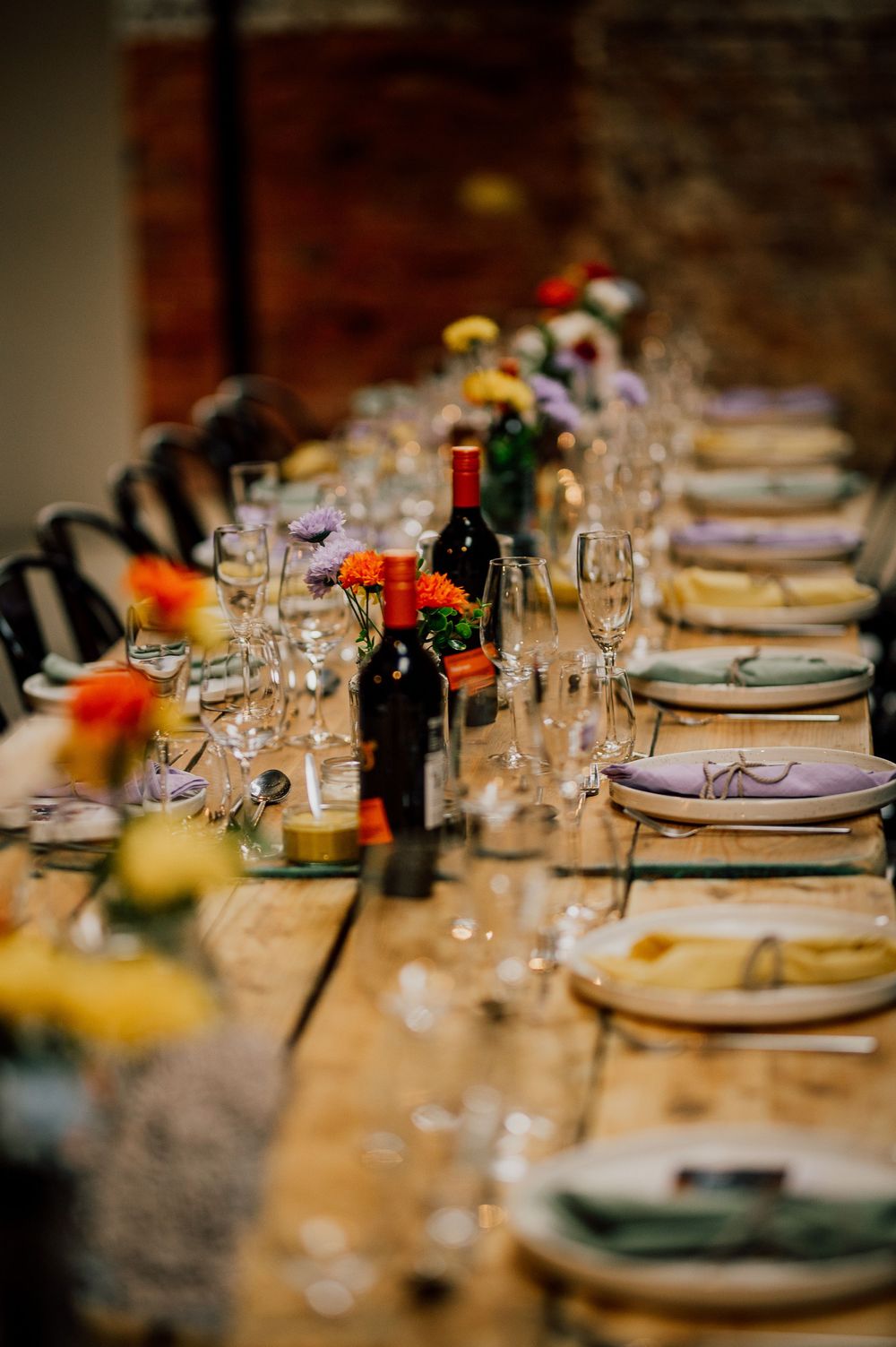 A rustic wooden table set for dinner with wine bottles, glasses and colorful floral accents.