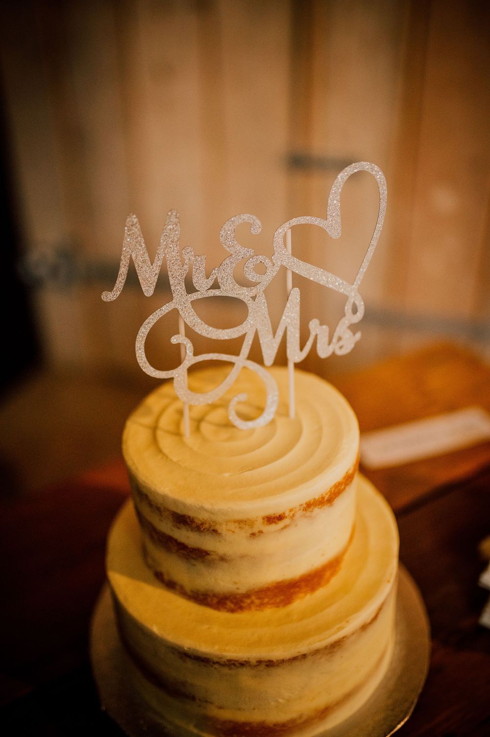 A three-tiered semi-naked wedding cake with white frosting and a glittery 'Mr & Mrs' cake topper with heart decoration.
