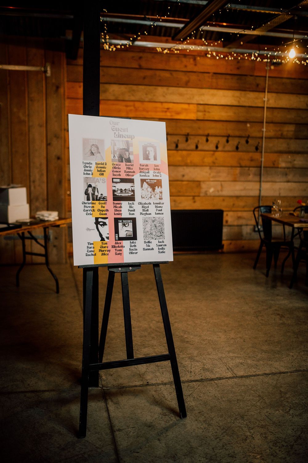 A wedding seating chart displayed on an easel in a rustic wooden venue with string lights overhead.