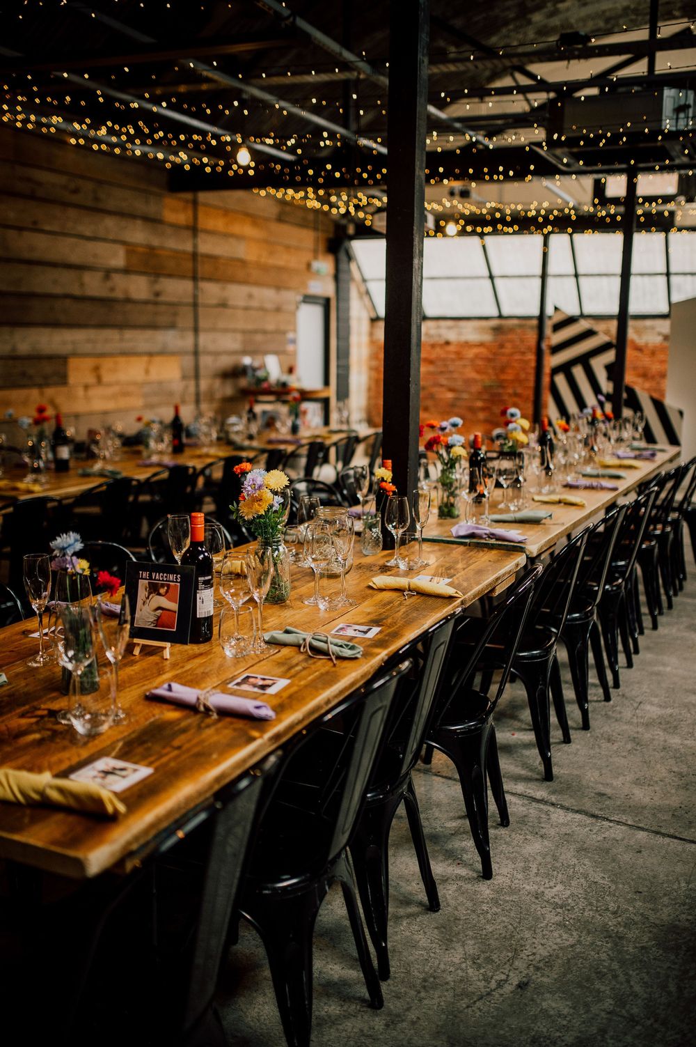 Long wooden dining table with black metal chairs set for dinner under string lights in rustic industrial venue.