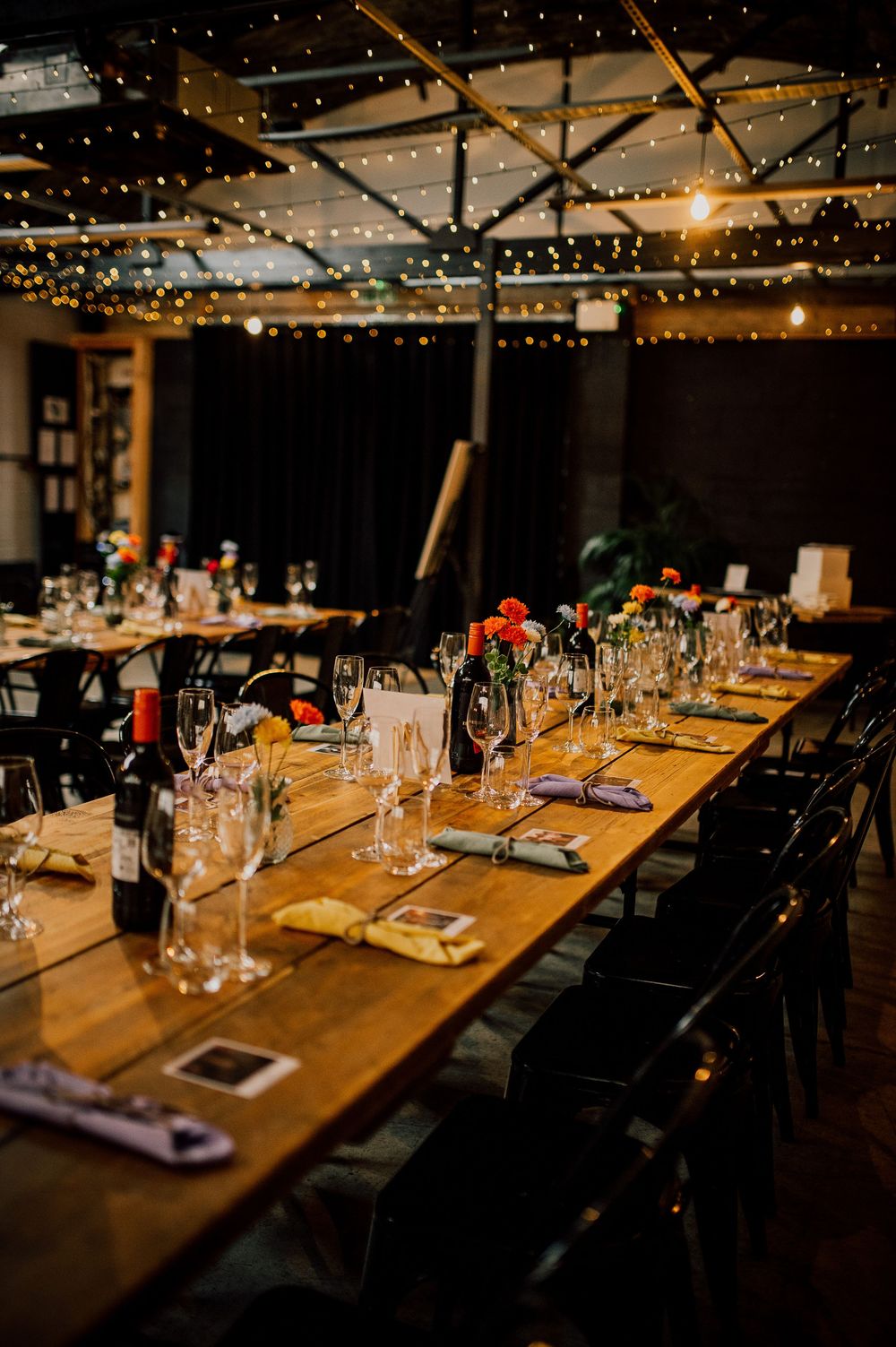 A rustic dining table set for an elegant dinner party with string lights overhead and wine glasses at each place setting.