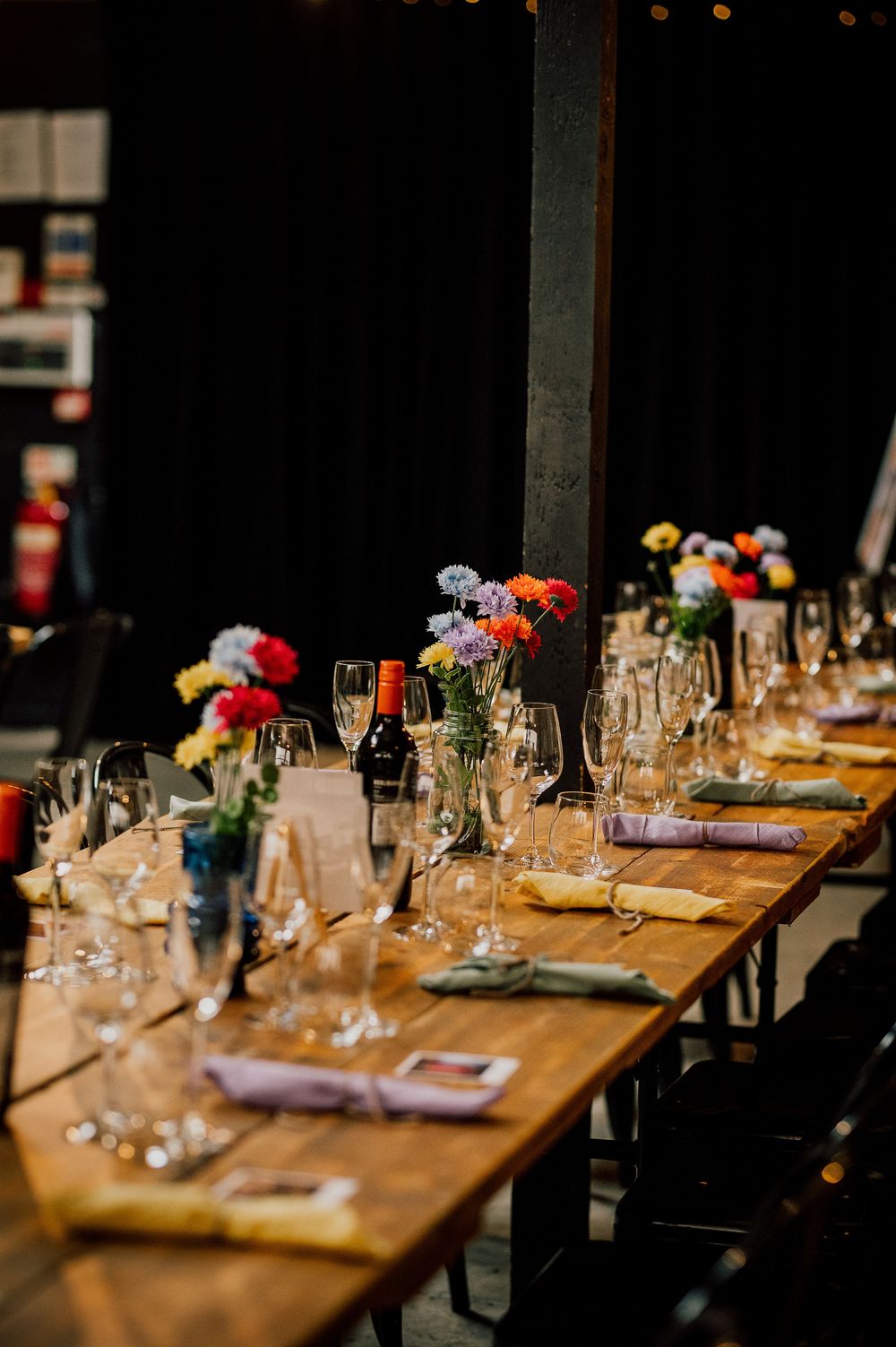 Elegant table setting with wooden tables, wine glasses, and colorful floral centerpieces in a dimly lit restaurant.