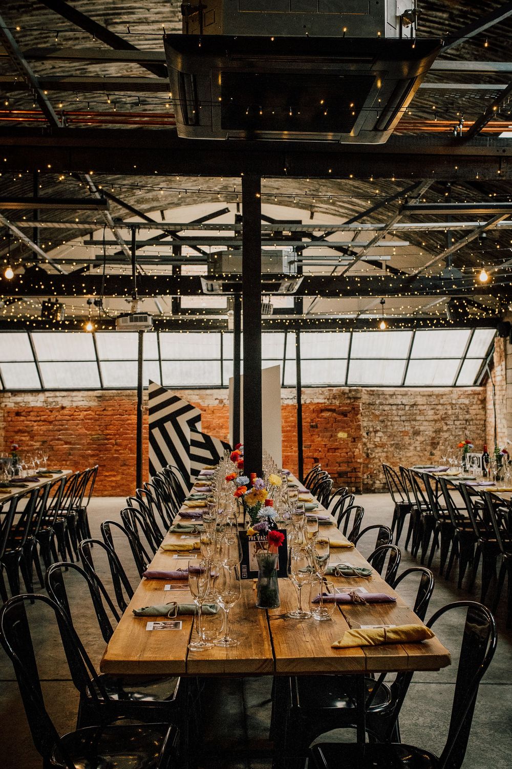 Rustic restaurant interior with exposed brick walls, wooden tables and black chairs arranged in long rows under string lights.