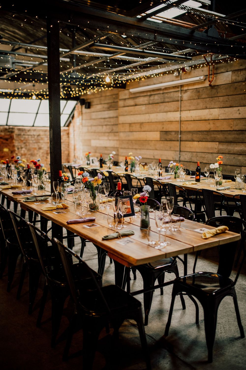 Long wooden tables with black chairs set for dinner under string lights in rustic industrial wedding venue.