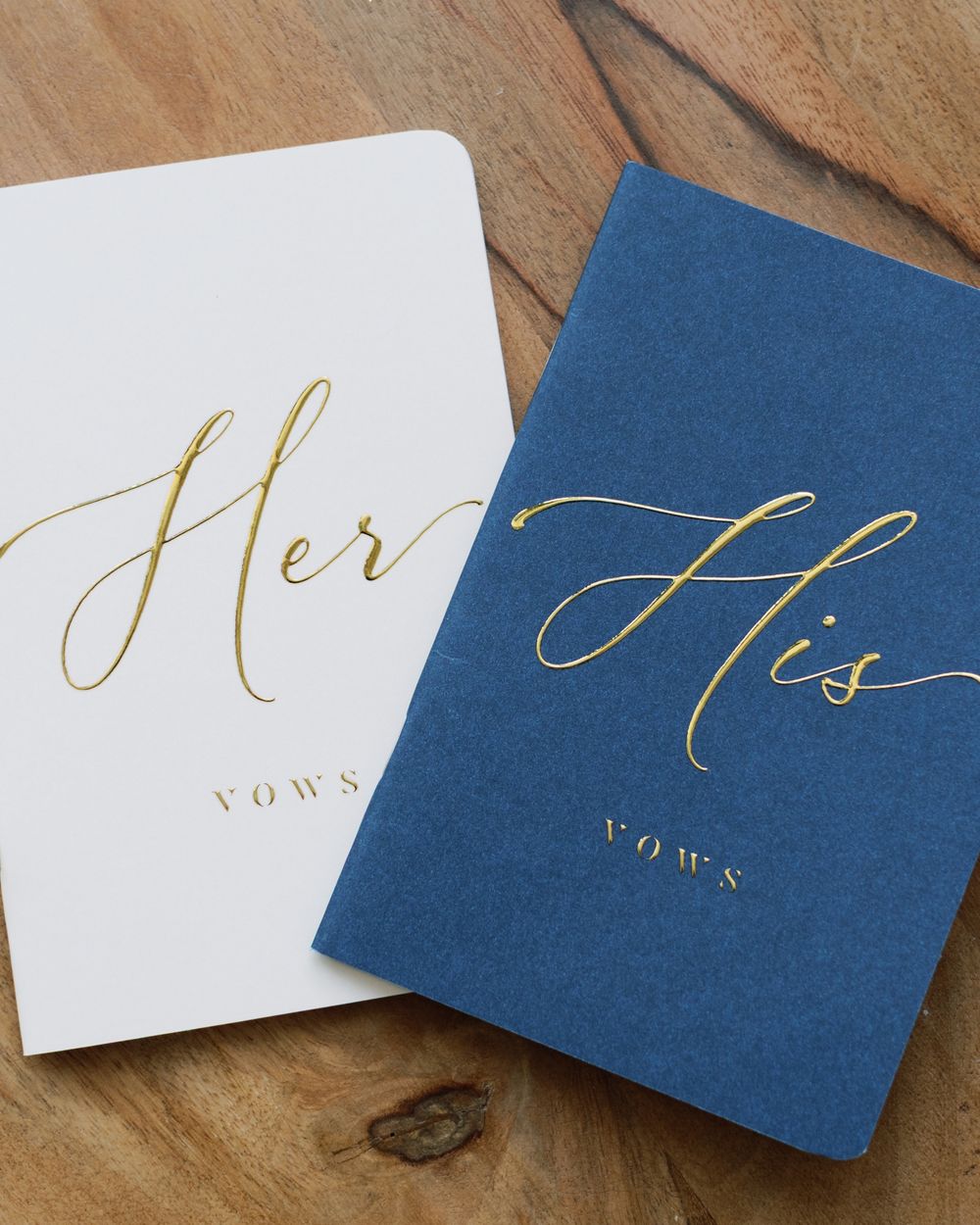 Elegant navy blue and white wedding vow books with gold calligraphy lettering on a wooden surface.