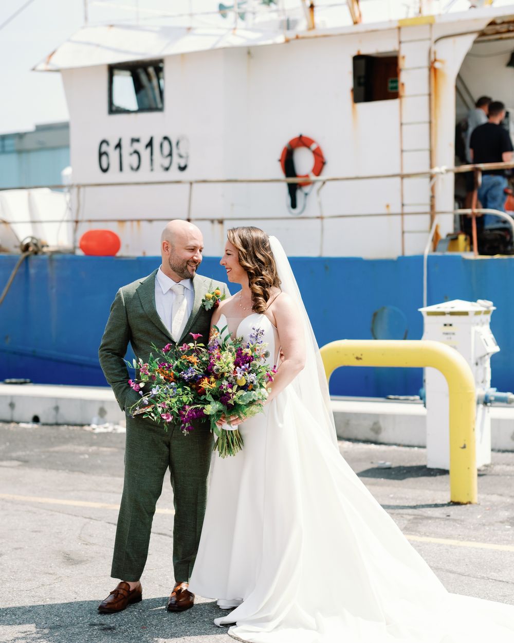 A couple in wedding attire share a romantic moment by a docked boat on a sunny day.