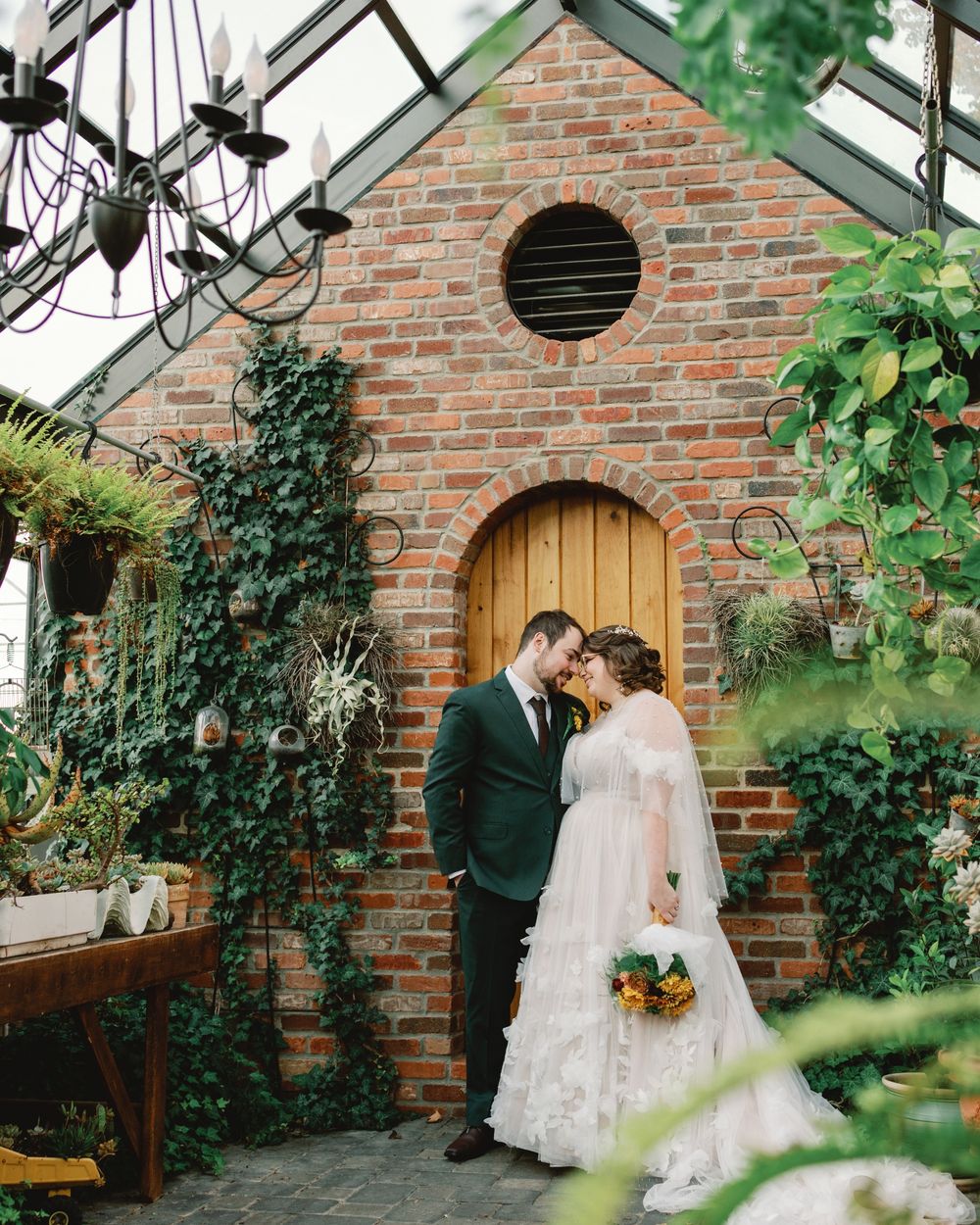 Romantic wedding couple embraces in front of a rustic brick greenhouse surrounded by lush greenery and climbing vines.