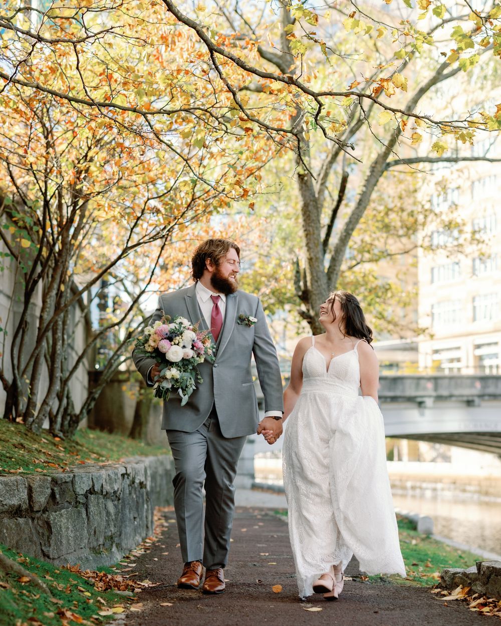 A couple walks along a tree-lined stone path during autumn with fallen leaves and warm golden light.