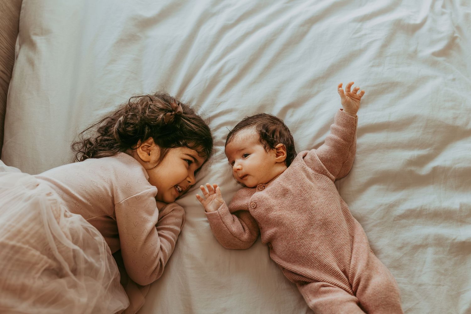 Two children sleeping peacefully together on white bedding while wearing matching pink sweaters.