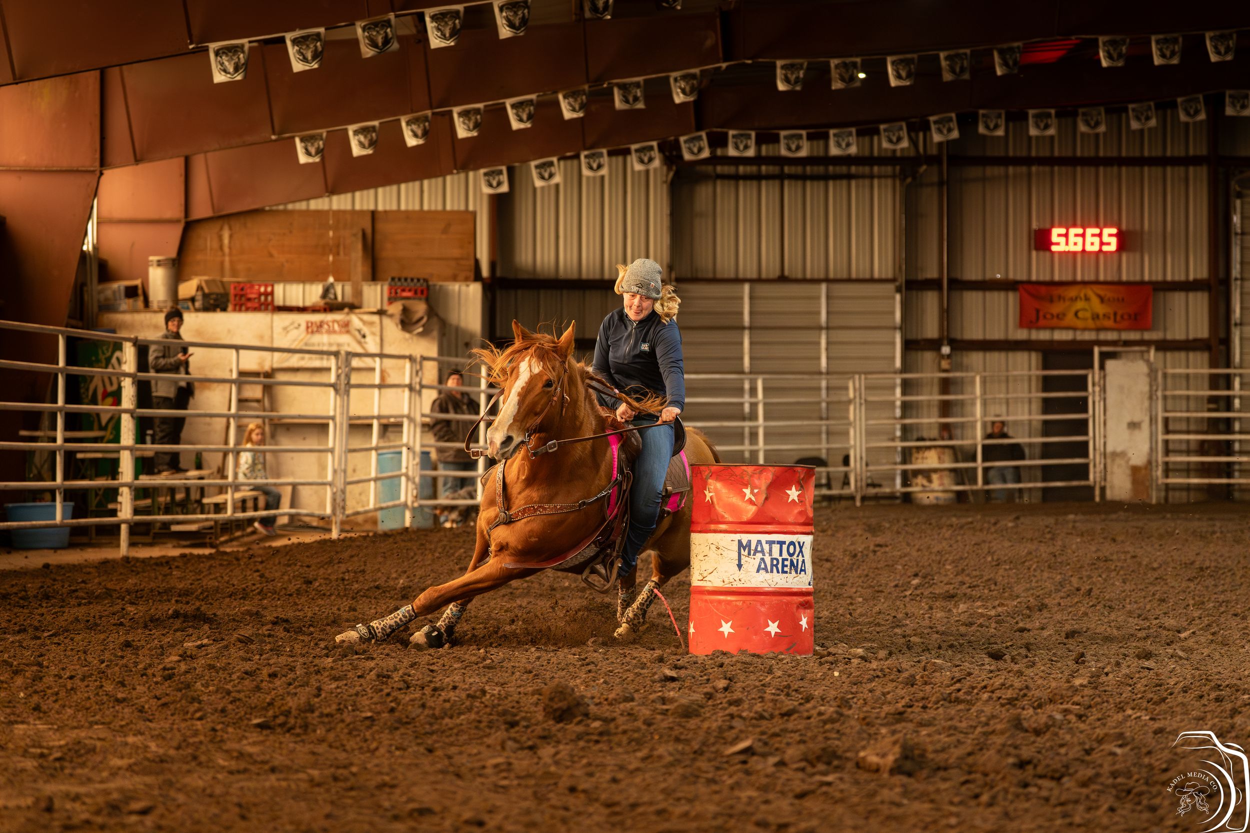 Equestrian athlete and horse navigating barrel racing turn in dimly lit indoor arena.