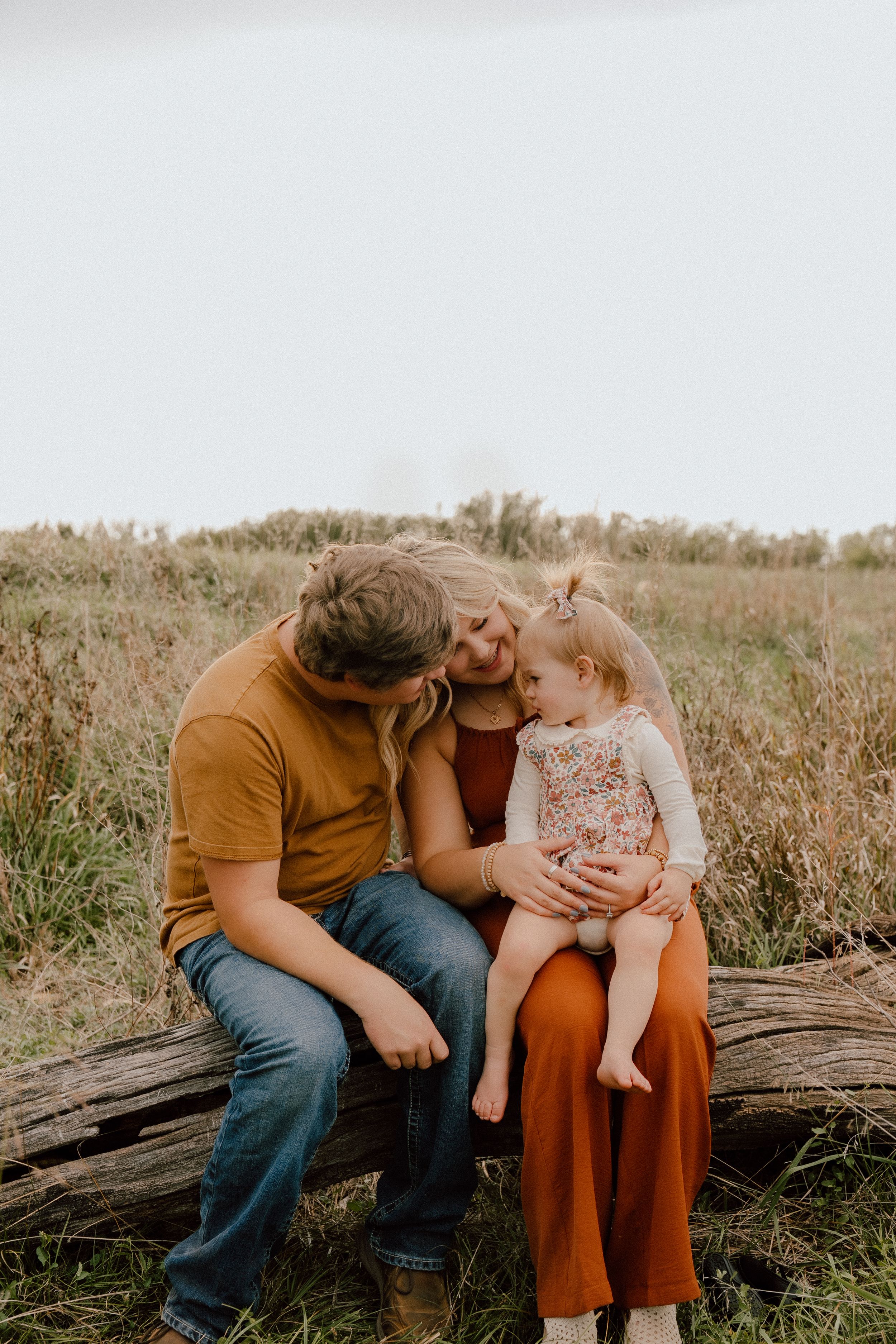 Young family of three sitting on a log in a golden meadow, sharing a tender moment outdoors.