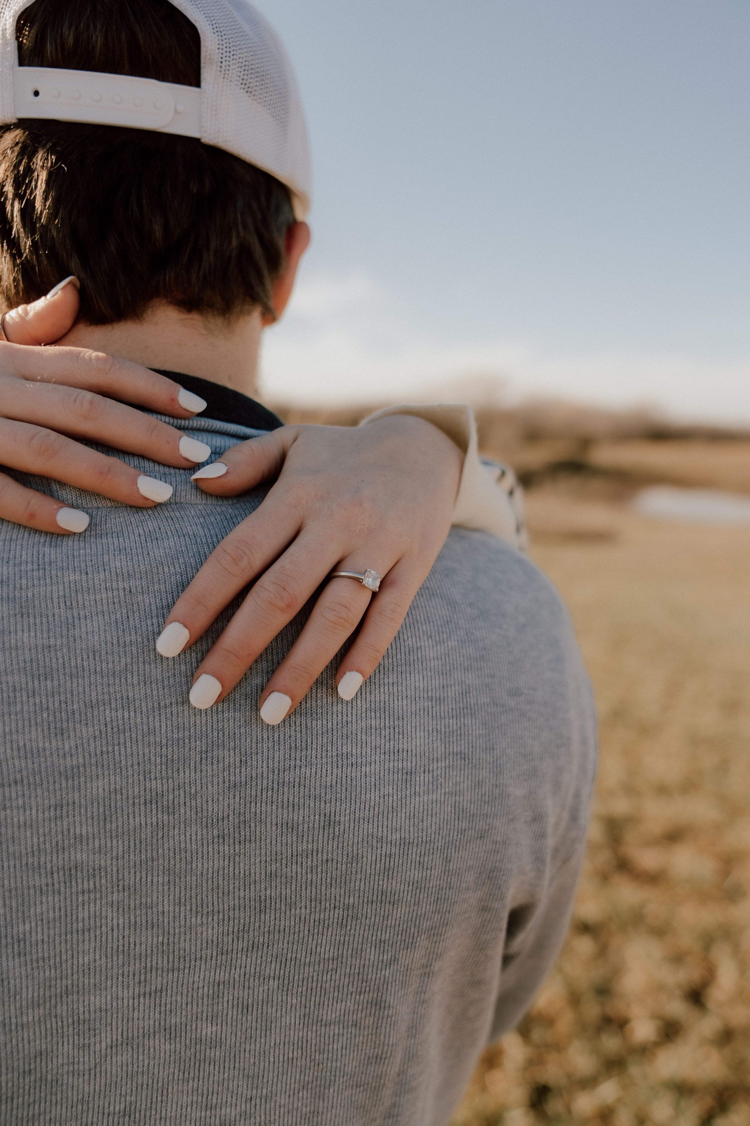 Close-up of woman's hand with engagement ring resting on man's shoulder in gray shirt outdoors.