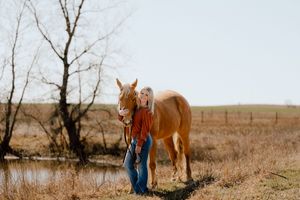Blonde woman standing with palomino horse in golden autumn field near pond.