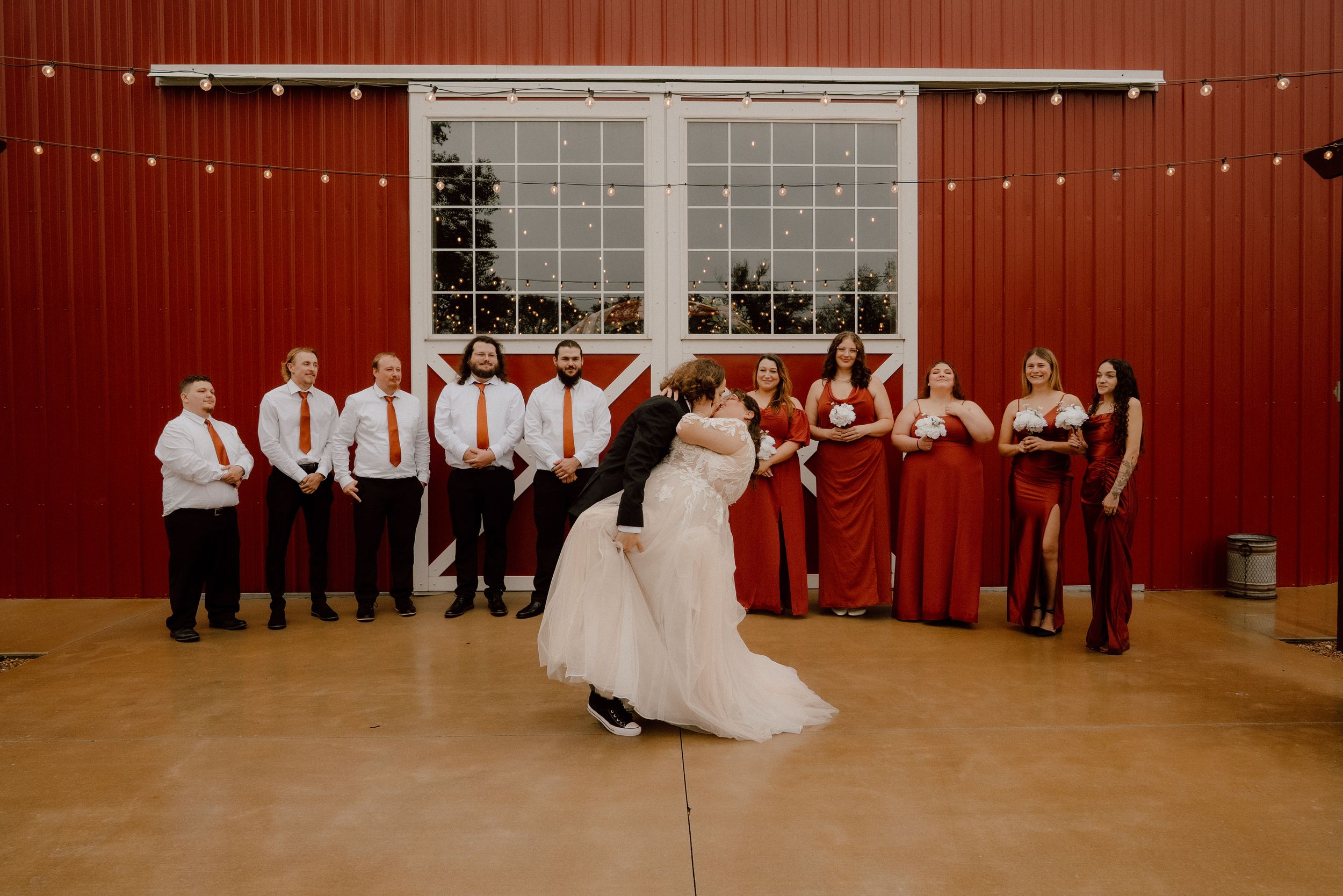 Bride and groom share first dance in red barn venue surrounded by wedding party in red attire.