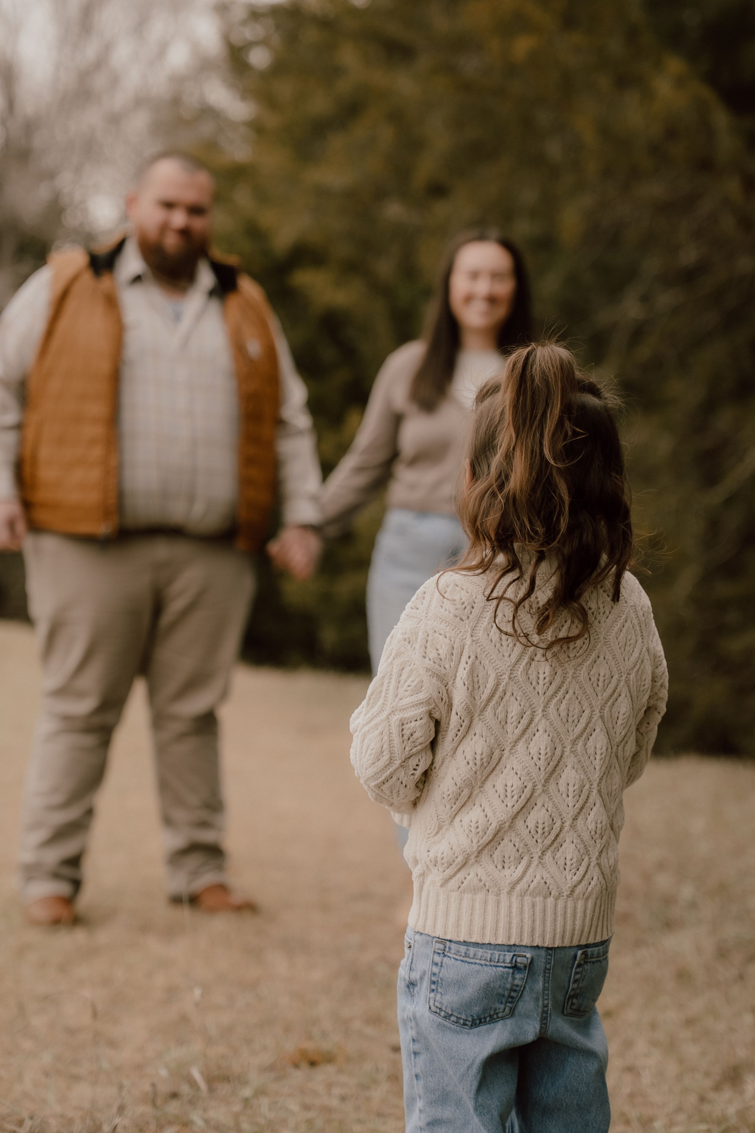 Young girl in cream sweater watches her parents hold hands in a blurred outdoor background.