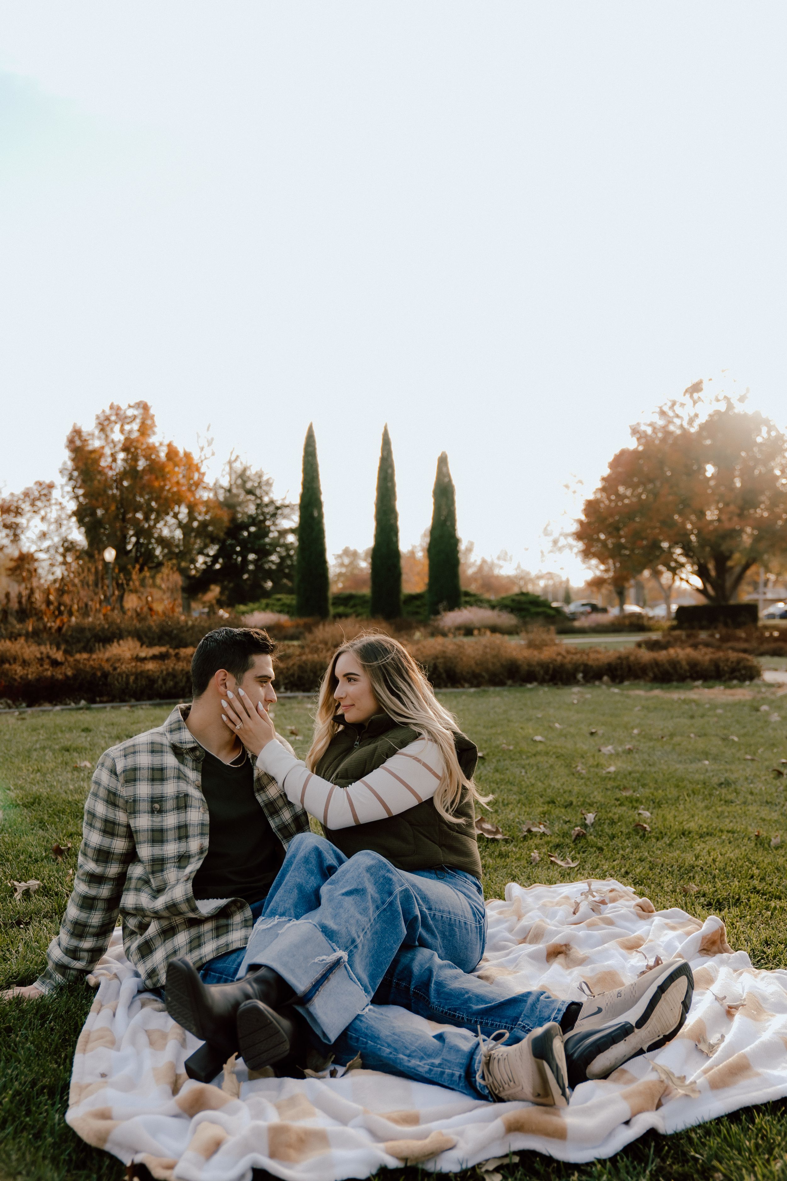 Couple sitting on a blanket in a park during fall, sharing a tender moment with autumn trees in the background.