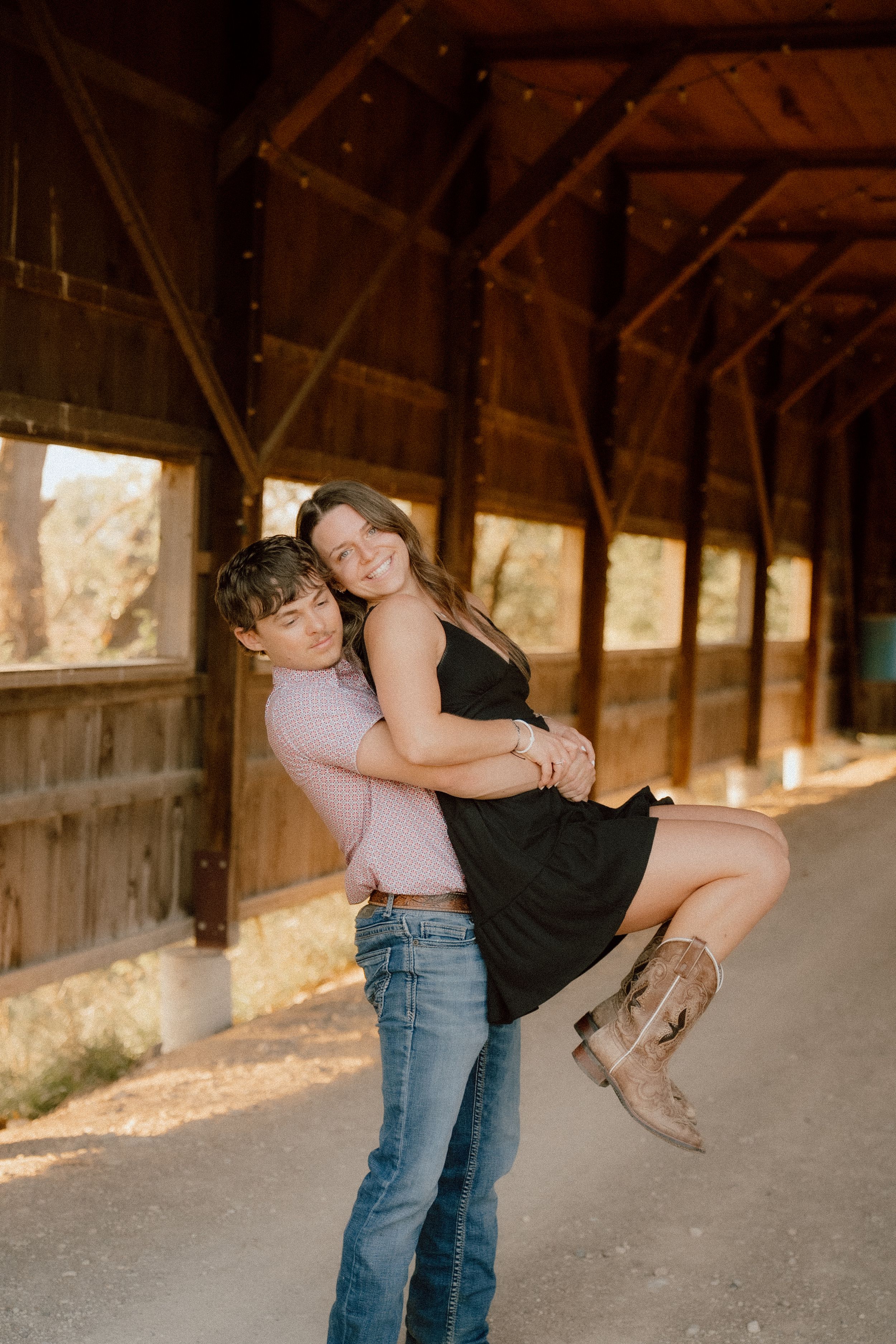 Man lifts smiling woman in black dress inside a rustic wooden barn during engagement photo session.