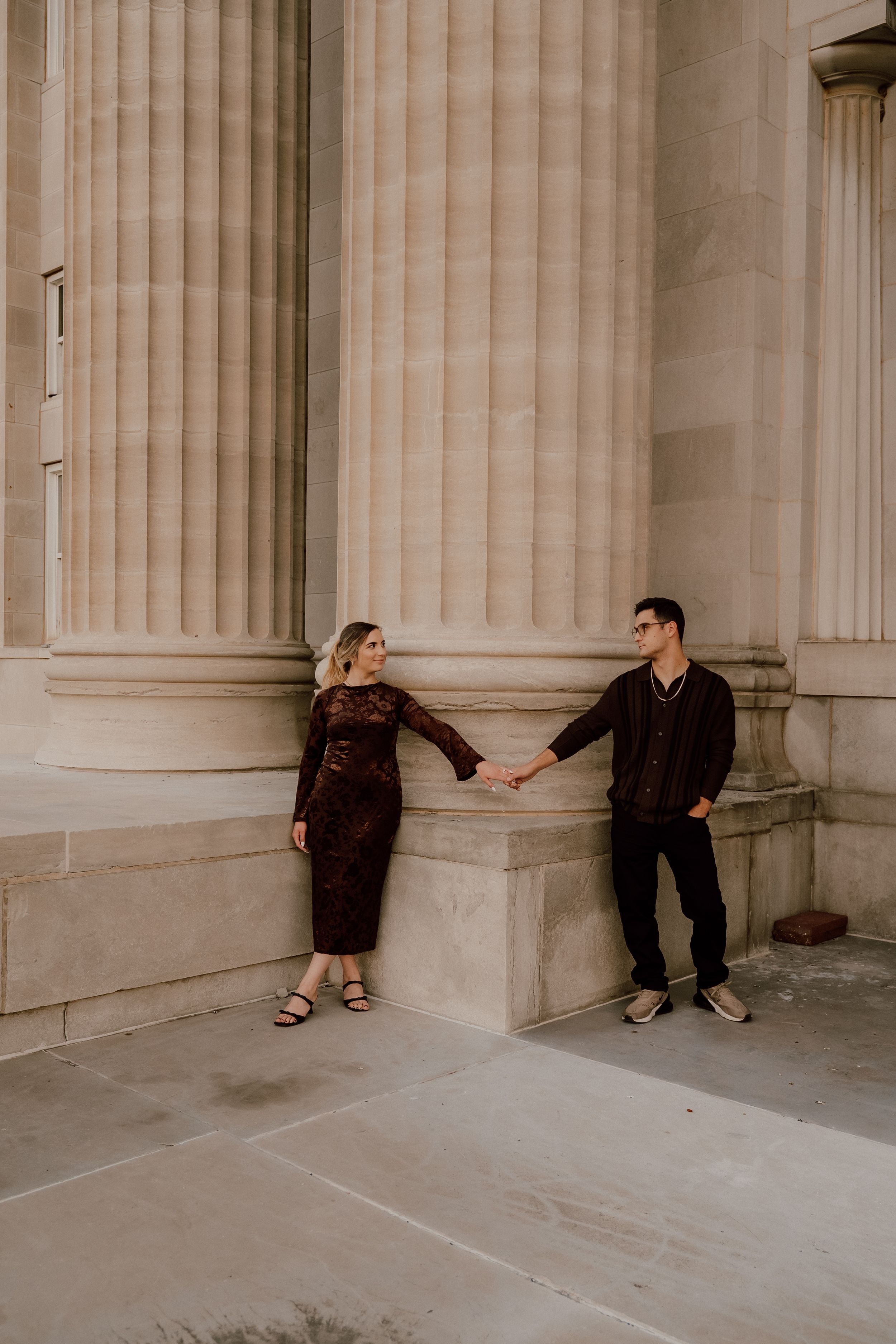 Couple holding hands between stone columns in matching dark brown outfits during an urban engagement photo session.