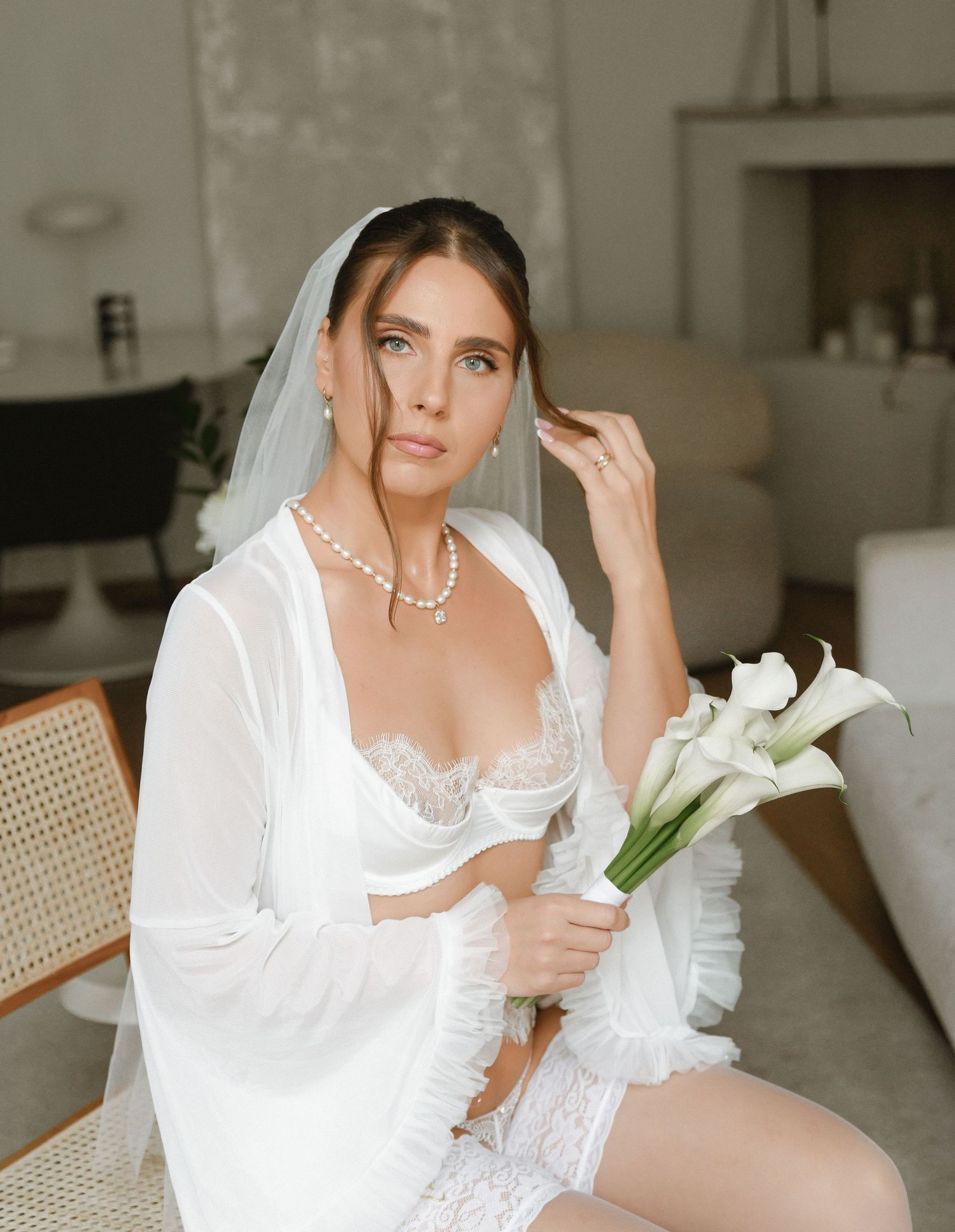 Elegant bridal boudoir photo featuring white lace lingerie, sheer robe, pearl necklace and calla lily bouquet.