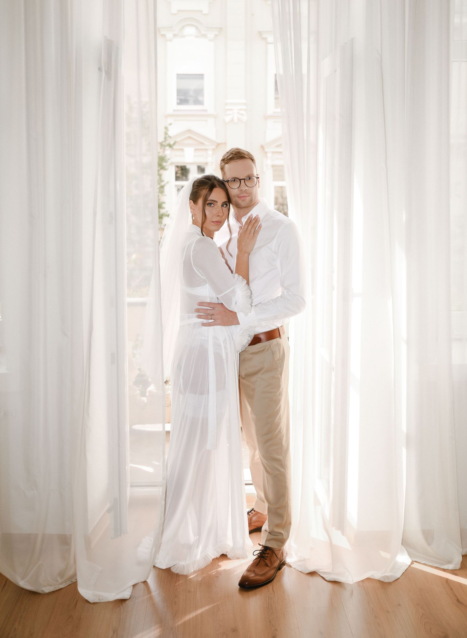 Romantic couple embracing by sheer white curtains in a sunlit room with hardwood floors.
