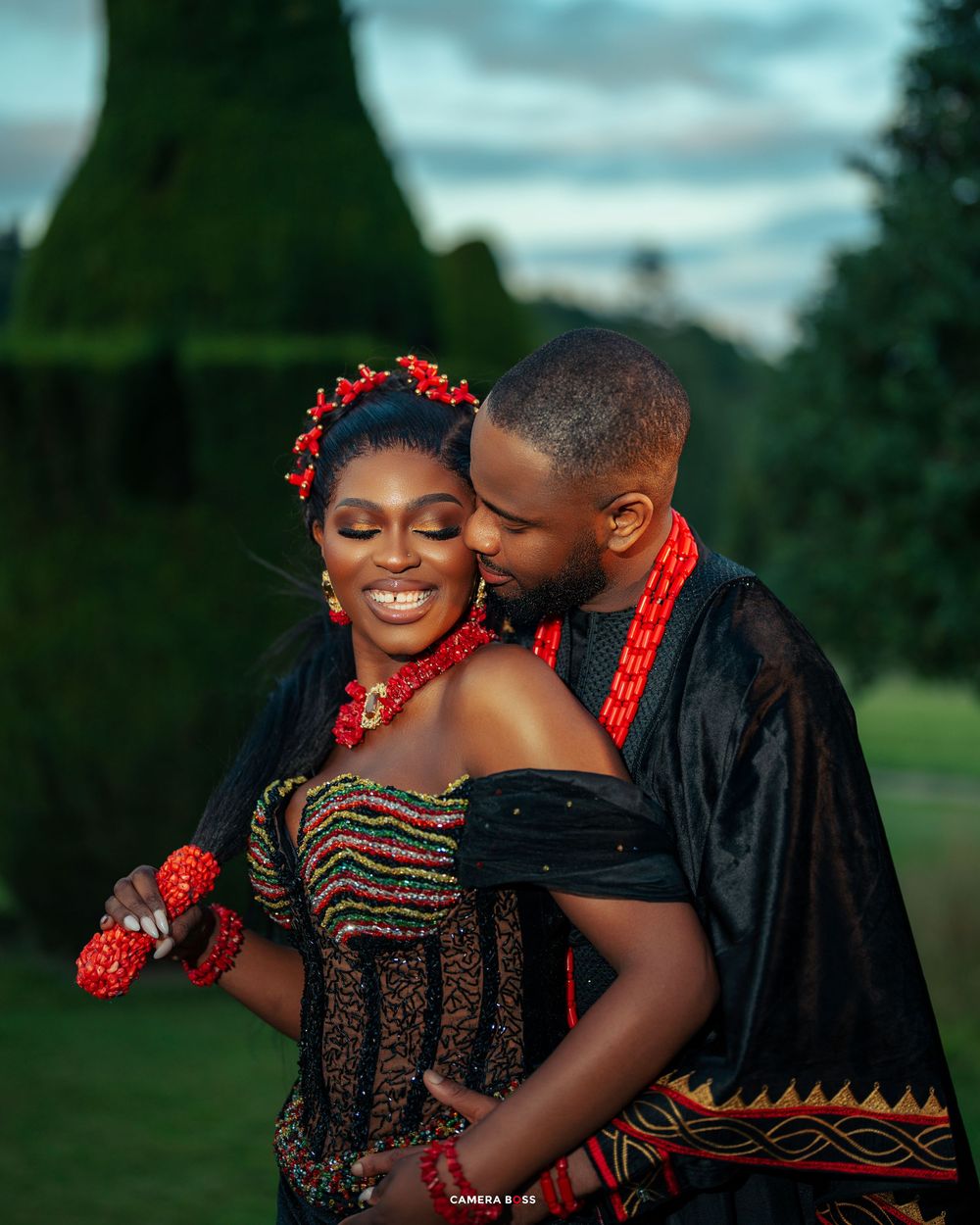 A couple in traditional African attire share romantic moments in a garden setting with red floral accessories.