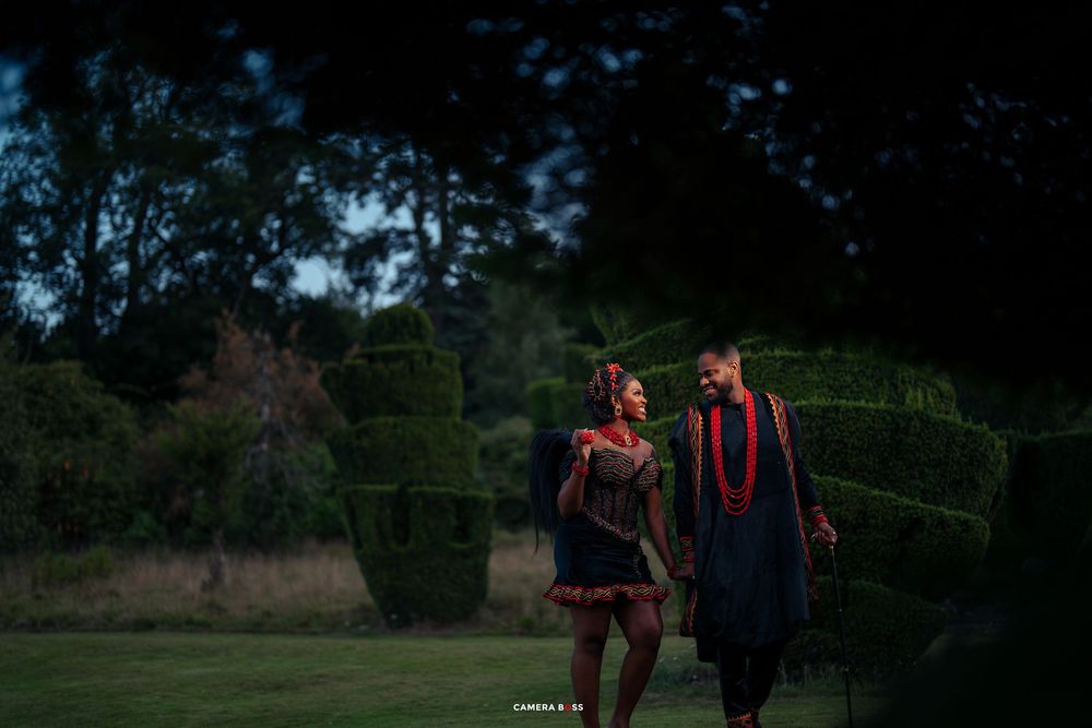 Two graduates in red and blue graduation gowns walk together in a garden with ornamental hedges at dusk.