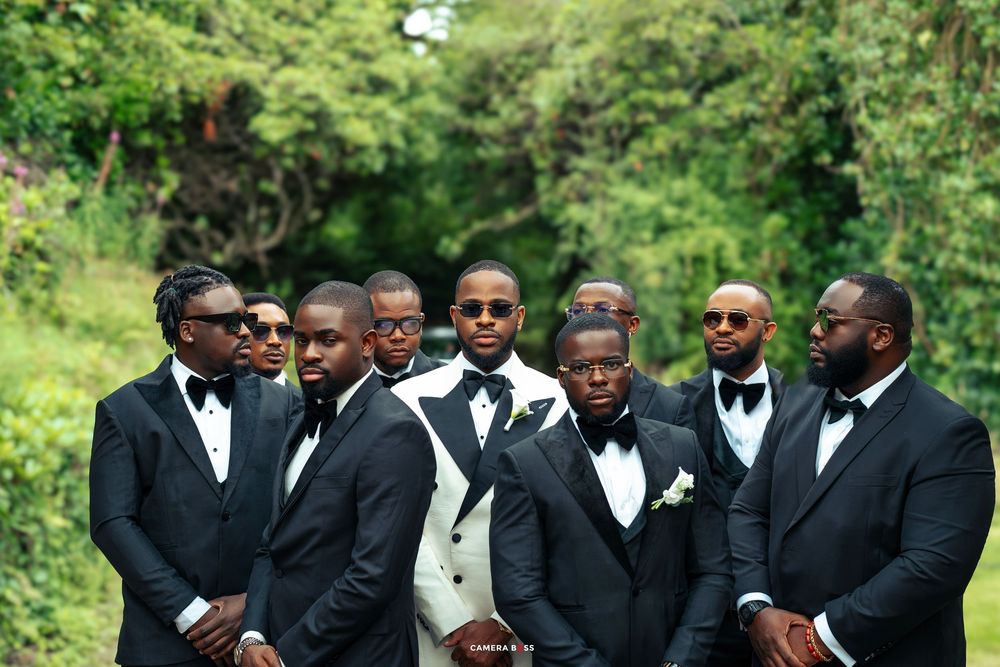 Groomsmen in black tuxedos and bow ties pose together outdoors during a wedding celebration.