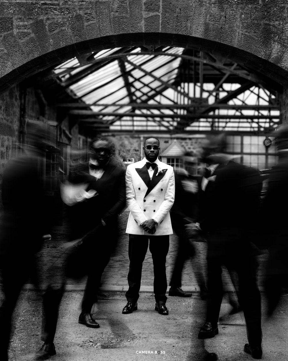 A black and white motion blur photograph shows figures moving through an industrial space with skylights overhead.