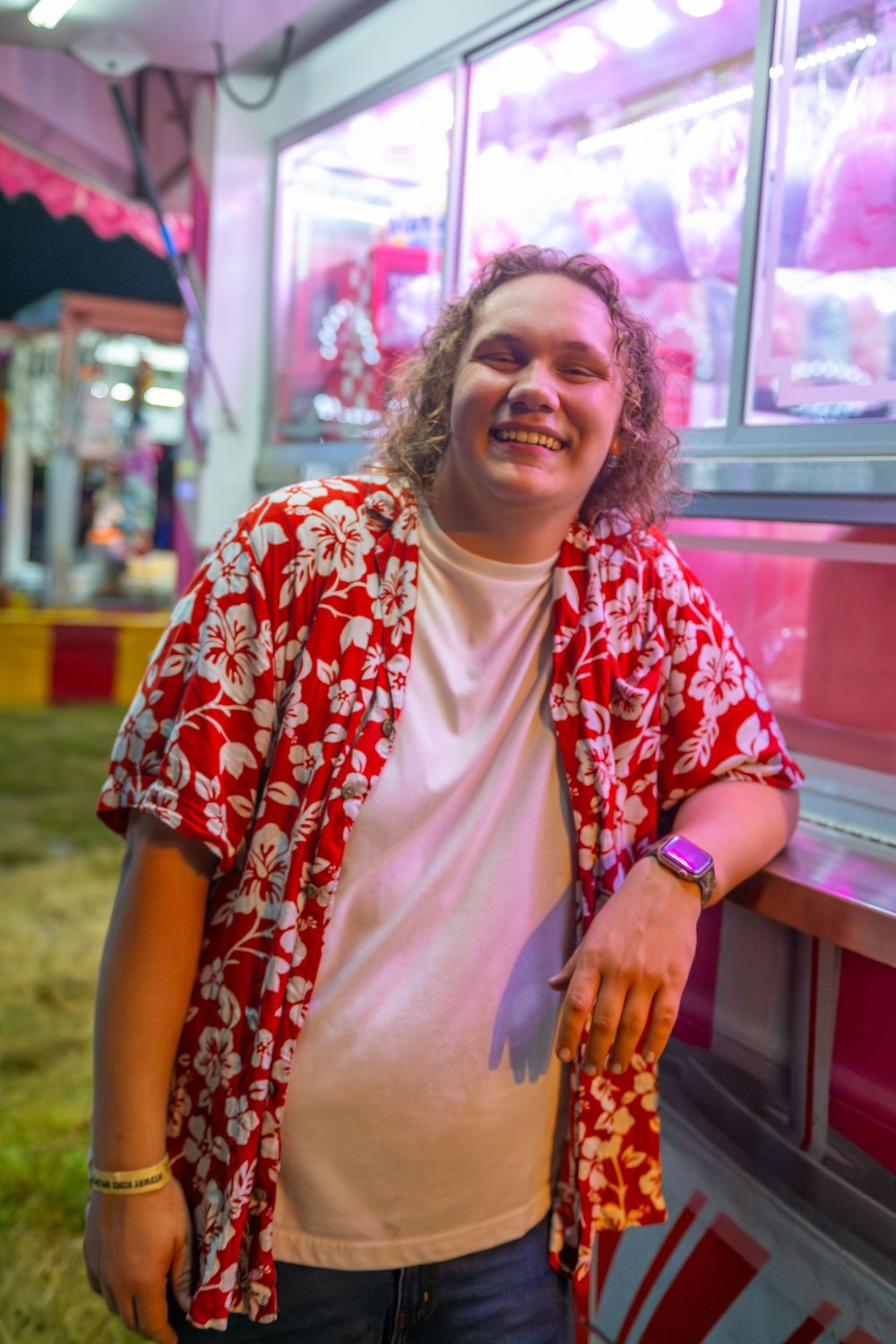 A person wearing a red floral shirt and white t-shirt smiles while standing in front of a pink-lit display case.